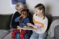 Eine Seniorin und zwei Kinder genießen das Lesen eines Buches auf einer Couch und lachen dabei herzlich miteinander.